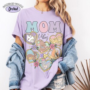 Pu&ograve; includere: Una maglietta lilla chiaro con le parole "MOM Vibes" in un font retr&ograve;. La maglietta presenta immagini di cartoni animati di personaggi Disney in cornici a forma di cuore. L'angolo in alto a sinistra della maglietta riporta le parole "COMFORT COLOR Orchid".