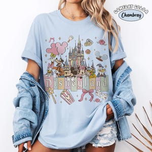 Peut inclure: T-shirt bleu clair avec un motif Disneyland. Le design pr&eacute;sente un ch&acirc;teau, Mickey Mouse, Minnie Mouse, Donald Duck, Dingo et d'autres personnages. Le mot "Disneyland" est &eacute;crit en couleurs pastel. Le t-shirt a des manches retrouss&eacute;es.