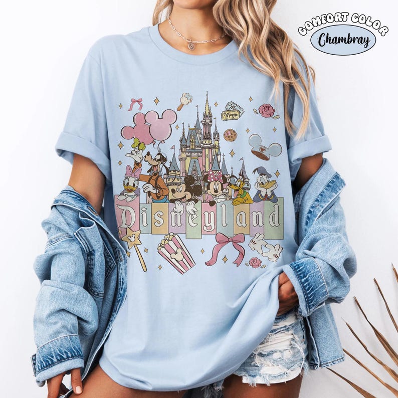 Peut inclure: T-shirt bleu clair avec un motif Disneyland. Le design pr&eacute;sente un ch&acirc;teau, Mickey Mouse, Minnie Mouse, Donald Duck, Dingo et d'autres personnages. Le mot "Disneyland" est &eacute;crit en couleurs pastel. Le t-shirt a des manches retrouss&eacute;es.
