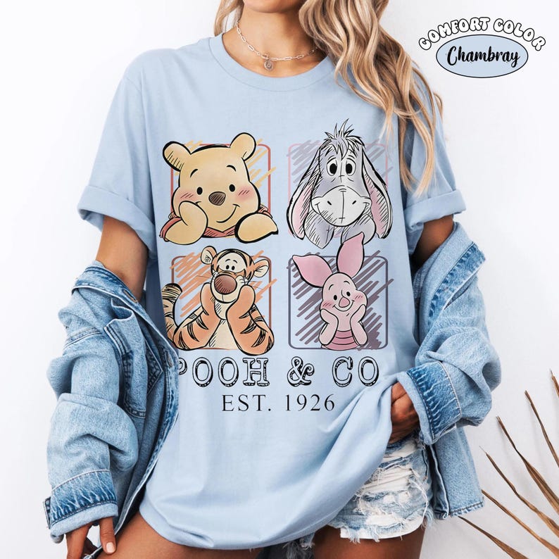 May include: Light blue Comfort Colors Chambray t-shirt featuring a graphic of Winnie the Pooh, Eeyore, Tigger, and Piglet. The design includes the text "Pooh & Co. EST. 1926". The shirt has rolled-up sleeves and is worn with a denim jacket.