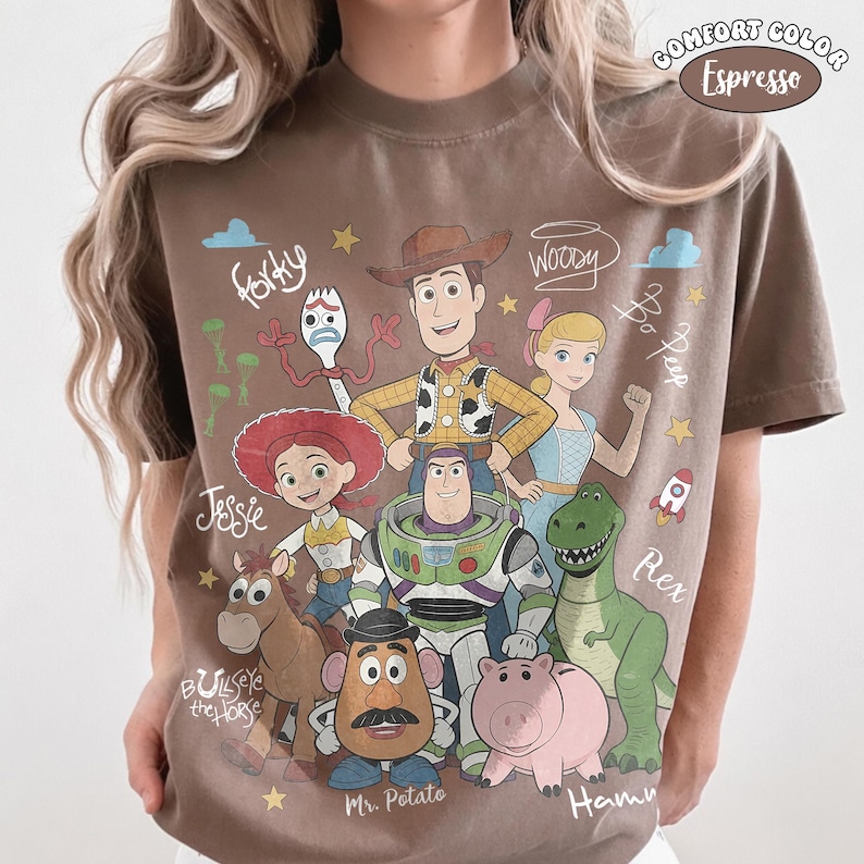 Może przedstawiać: Koszulka w kolorze espresso z nadrukiem graficznym przedstawiającym postacie z Toy Story, w tym Chudego, Buzza Astrala, Jessie, Celka, Rexa, Hamm, Pana Bulwę, Pastereczkę i Sztućka, z odręcznie napisanymi imionami oraz małymi gwiazdkami i chmurkami.