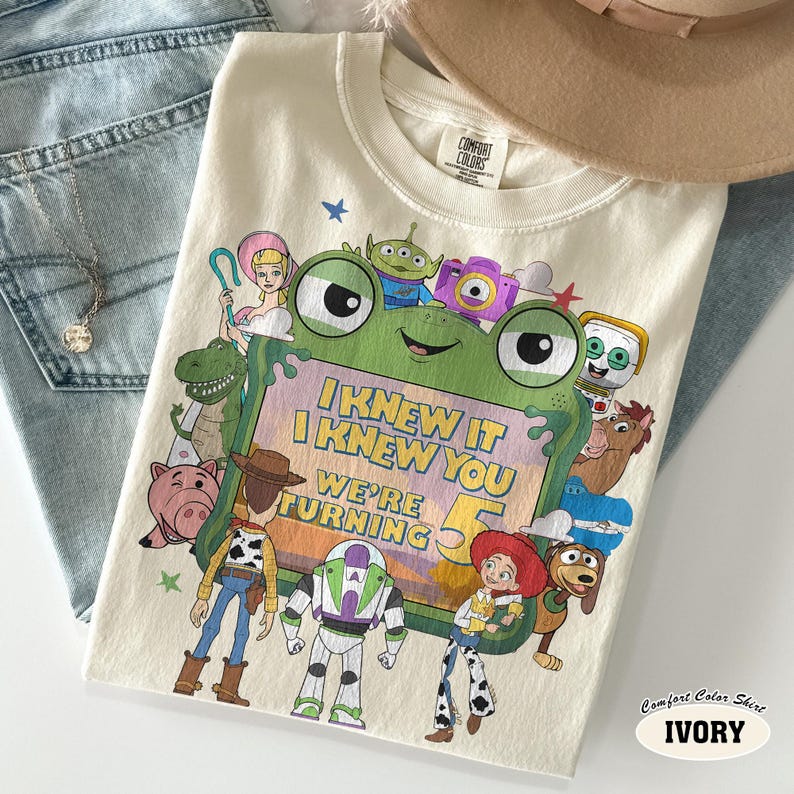 Puede incluir: Una camiseta de color marfil con un gran marco verde en forma de rana que contiene el texto I KNEW IT I KNEW YOU WE'RE TURNING 5. El marco est&aacute; rodeado por varios personajes animados, incluyendo un vaquero, un guardi&aacute;n espacial, una vaquera y un dinosaurio.