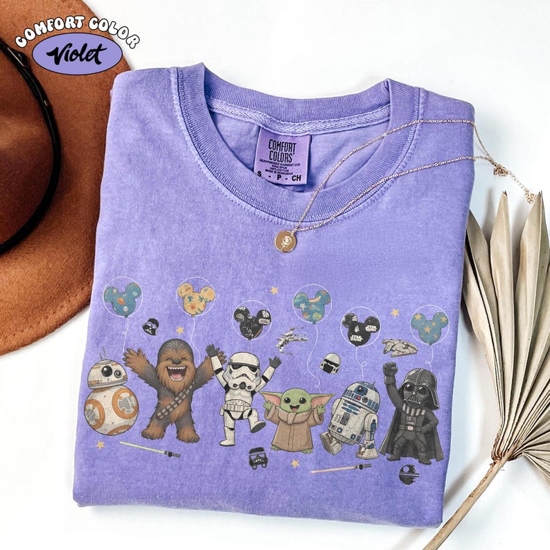 May include: Violet Comfort Colors t-shirt featuring Star Wars characters and Mickey Mouse-shaped balloons. The design includes BB-8, Chewbacca, a Stormtrooper, Grogu, R2-D2, and Darth Vader. A gold necklace and a brown hat are also visible.