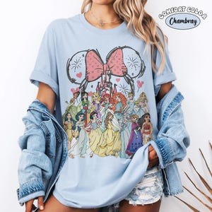 Op de afbeelding: Lichtblauw Comfort Colors Chambray T-shirt met een afbeelding van Disney-prinsessen, een roze strik en een kasteel. Het ontwerp bevat vuurwerk en hartaccenten. Het shirt heeft een relaxte pasvorm.