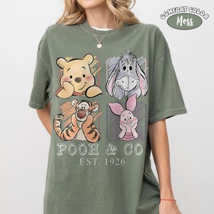 May include: Comfort Colors moss green t-shirt featuring a graphic of Winnie the Pooh, Eeyore, Tigger, and Piglet. The shirt has the text "POOH & CO EST. 1926" in white. The shirt is a short-sleeved crew neck.
