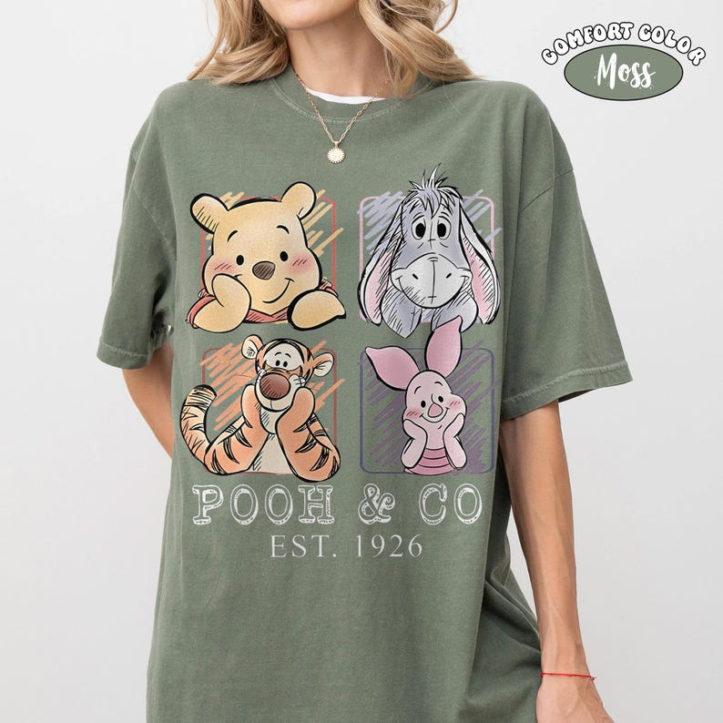 May include: Comfort Colors moss green t-shirt featuring a graphic of Winnie the Pooh, Eeyore, Tigger, and Piglet. The shirt has the text "POOH & CO EST. 1926" in white. The shirt is a short-sleeved crew neck.