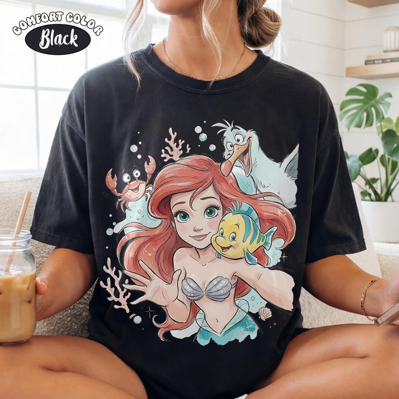 May include: Black Comfort Colors t-shirt with a vibrant illustration of Ariel, Flounder, Sebastian, and Scuttle from The Little Mermaid. The design includes coral and bubbles, with the text "Comfort Color Black" in a white oval.