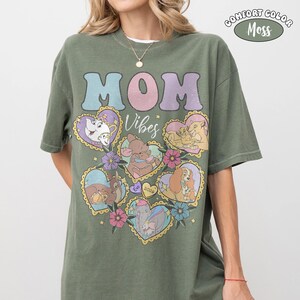 Pu&ograve; includere: T-shirt verde oliva con le parole "MOM Vibes" in colori pastello. La maglietta presenta illustrazioni a forma di cuore di personaggi Disney. L'angolo in alto a destra ha un logo "Comfort Color Moss".
