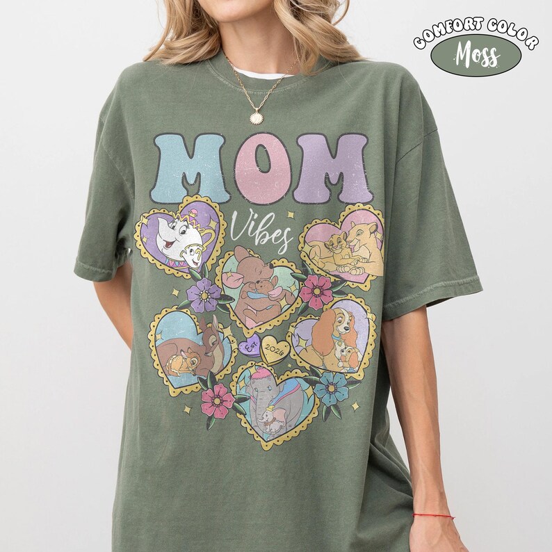 Pu&ograve; includere: T-shirt verde oliva con le parole "MOM Vibes" in colori pastello. La maglietta presenta illustrazioni a forma di cuore di personaggi Disney. L'angolo in alto a destra ha un logo "Comfort Color Moss".