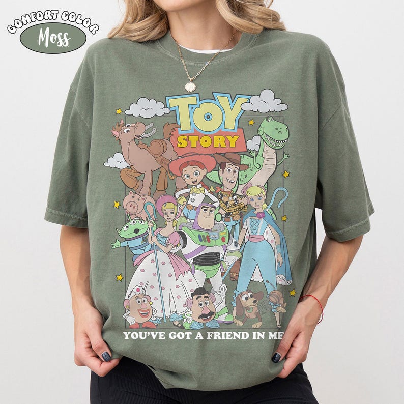 K&ouml;nnte beinhalten: Ein moosgr&uuml;nes T-Shirt mit einem Grafikdruck der Toy Story-Figuren, darunter Woody, Buzz Lightyear und Jessie. Der Text lautet TOY STORY und YOU'VE GOT A FRIEND IN ME. Das T-Shirt hat kurze &Auml;rmel und einen Rundhalsausschnitt.