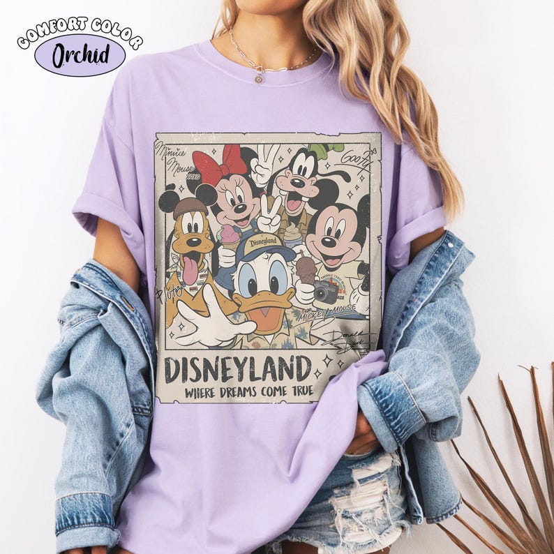 Puede incluir: Una camiseta de color morado claro con un estampado gr&aacute;fico rectangular de personajes de Disney como Mickey Mouse, Minnie Mouse, el Pato Donald, Goofy y Pluto. El texto dice Disneyland Where Dreams Come True, con nombres en cursiva.