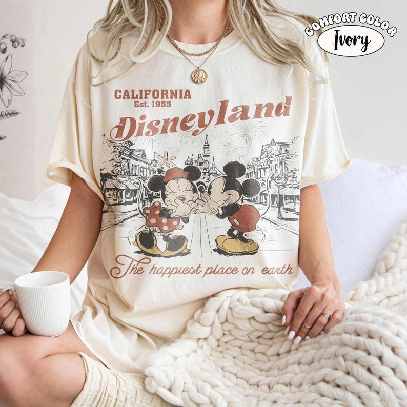 May include: An ivory short-sleeve t-shirt featuring a brown graphic print of Mickey and Minnie Mouse in front of a castle. The text reads California Est. 1955, Disneyland, and The happiest place on earth. A small circular logo says Comfort Color Ivory.