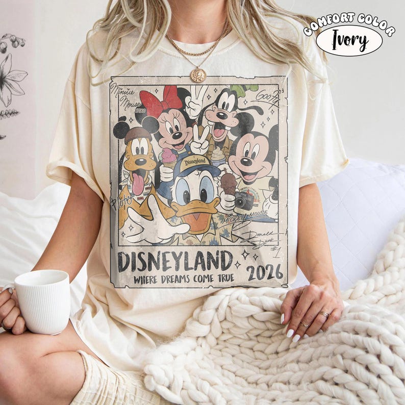 Puede incluir: Una camiseta de manga corta color marfil con un estampado gr&aacute;fico rectangular de personajes de Disney, incluidos Mickey Mouse, Minnie Mouse, el Pato Donald, Goofy y Pluto. El texto dice Disneyland, Where Dreams Come True y 2026.