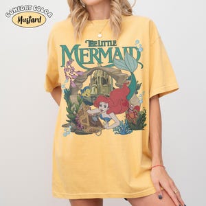 K&ouml;nnte beinhalten: Senffarbenes T-Shirt mit einer Grafik von Arielle aus Arielle, die Meerjungfrau. Das Design zeigt Arielle, Fabius, Sebastian und ein Schloss. Das Shirt tr&auml;gt den gr&uuml;nen Schriftzug "The Little Mermaid".