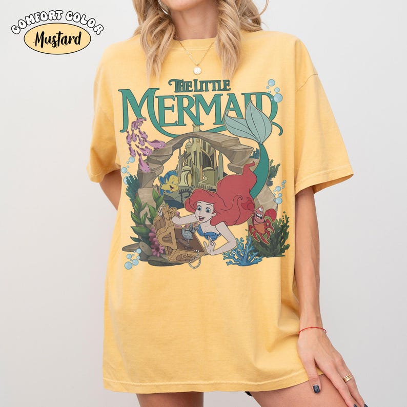 K&ouml;nnte beinhalten: Senffarbenes T-Shirt mit einer Grafik von Arielle aus Arielle, die Meerjungfrau. Das Design zeigt Arielle, Fabius, Sebastian und ein Schloss. Das Shirt tr&auml;gt den gr&uuml;nen Schriftzug "The Little Mermaid".