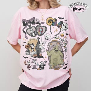 May include: A light pink t-shirt featuring a graphic print of characters and icons from The Nightmare Before Christmas, including Jack Skellington, Sally, Zero, and Oogie Boogie, with a Comfort Color Blossom logo in the top right corner.