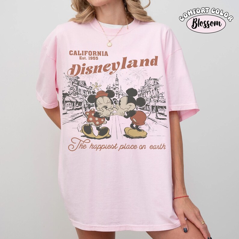 May include: A light pink short-sleeve t-shirt featuring a graphic print of Mickey and Minnie Mouse. The text reads California Est. 1955 Disneyland The happiest place on earth. A small circular logo in the top right corner says Comfort Color Blossom.
