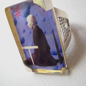 May include: A silver ring with a rectangular blue and gold resin inlay featuring a black and white image of a person in a robe. The resin inlay has a gold border and the word "禅" in red.