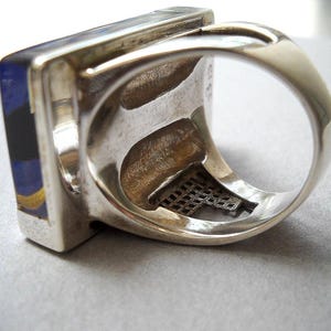 May include: A silver ring with a square blue, black, and gold stone set in the band. The ring has a decorative metal mesh design on the inside of the band.