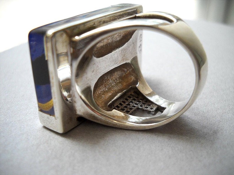 May include: A silver ring with a square blue, black, and gold stone set in the band. The ring has a decorative metal mesh design on the inside of the band.