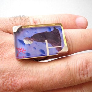 May include: A silver ring with a blue and white rectangular glass stone featuring a person in a black robe. The stone has a red inscription that says "生".