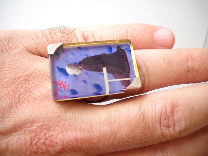 May include: A silver ring with a blue and white rectangular glass stone featuring a person in a black robe. The stone has a red inscription that says "生".
