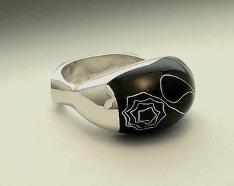May include: A silver ring with a black, rounded top. The black surface features a white, stylized floral design. The ring has a smooth, polished finish, reflecting light.