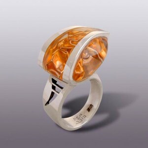 May include: A silver ring with a large, faceted, orange gemstone set in a curved, silver band. The band is marked with the text "GIFT 925".