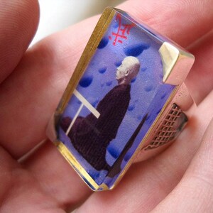 May include: A silver ring with a blue and gold resin inlay featuring a man in a dark suit. The resin inlay has a white background with blue and white clouds. The ring has a silver band with a textured design.