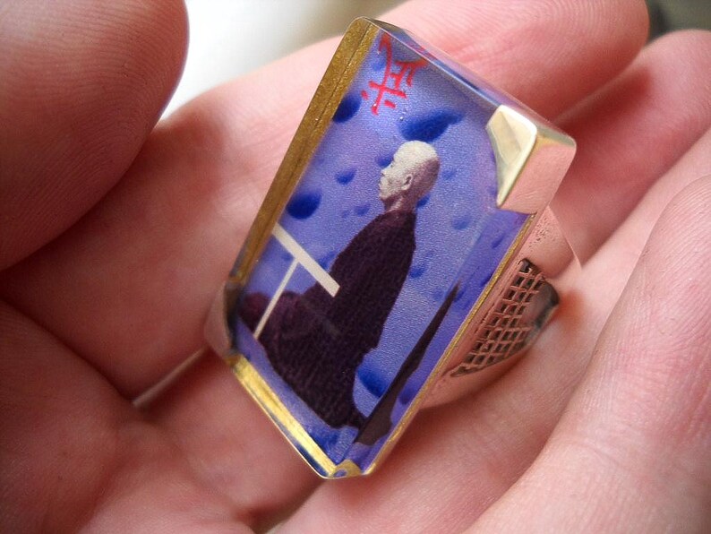 May include: A silver ring with a blue and gold resin inlay featuring a man in a dark suit. The resin inlay has a white background with blue and white clouds. The ring has a silver band with a textured design.