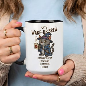 Puede incluir: Taza de cerámica blanca con interior y asa negros. La taza presenta el texto "Cat's Wake-Up Brew" y una ilustración de un gato con sombrero de mago sosteniendo una taza de café. Texto adicional enumera los beneficios.