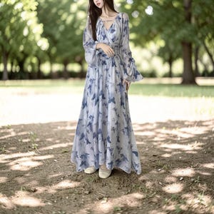 May include: A long, flowing dress with a butterfly print in shades of blue and white. The dress has long sleeves with flared cuffs and a V-neck. The model is wearing beige flats. The dress is made of a lightweight, sheer fabric.