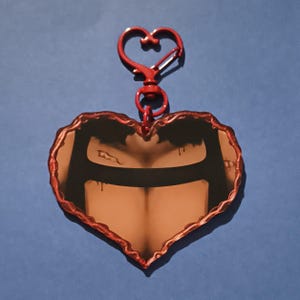 Peut inclure: Un porte-clés en forme de cœur avec une bordure et un clip rouges. L'image à l'intérieur du cœur représente un torse avec une bande noire sur la poitrine. Le fond est bleu uni.