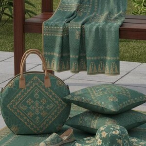 May include: An assortment of green and beige patterned items, including a tote bag, pillows, shoes, a sun hat, and a blanket. The items are arranged on a matching rug, creating a cohesive aesthetic. The scene is set outdoors, with a wooden bench and greenery in the background.