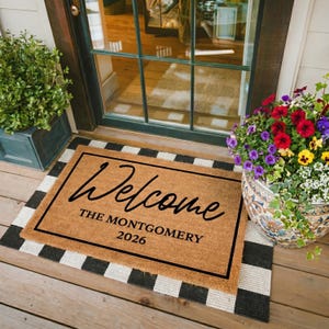 May include: A rectangular coir doormat with the word "Welcome" in black script, and the name "THE MONTGOMERY 2026" below. The mat is framed with a black border and sits on a black and white striped rug.