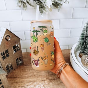 May include: A frosted glass tumbler with a bamboo lid, decorated with cartoon characters in various colors. The tumbler is held in a hand, with a metal house and Christmas decorations in the background.