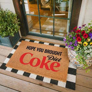 May include: A rectangular doormat with the words "HOPE YOU BROUGHT Coke Diet" in black and red. The mat has a natural brown background with a black and white striped border. The mat is placed in front of a door.