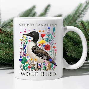 May include: White ceramic mug featuring a colorful illustration of a loon bird amidst flowers. The mug has the text "STUPID CANADIAN" above the bird and "WOLF BIRD" below. The handle is white.