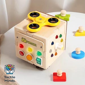 May include: A wooden sensory cube toy with a yellow fidget spinner on top. The cube features colorful buttons, a switch, and various shapes. The text "POWER" is visible on the front. Additional colorful shapes are scattered around the cube.
