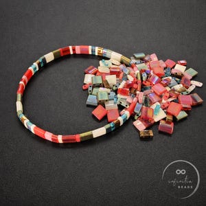May include: A beaded bracelet with a mix of red, white, and iridescent beads. A pile of loose, colorful beads in various shapes and sizes sits next to the bracelet. The beads are on a black surface.