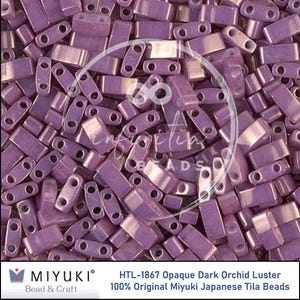 May include: Close-up of a pile of HTL-1867 Opaque Dark Orchid Luster Miyuki Japanese Tila Beads. The beads are a rectangular shape with two holes and a lustrous finish. The beads are a dark purple color.
