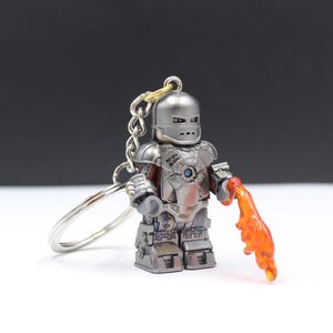 May include: A silver-colored armored character keychain featuring a metallic helmet, chest plate with a circular blue center, and blocky limbs. The figure holds a translucent orange flame-shaped accessory in one hand and is attached to a metal ring.