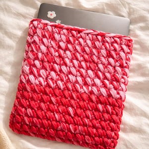 May include: A red and pink crocheted laptop sleeve. The sleeve is made of a thick, textured yarn and features a gradient design, transitioning from a deep red at the bottom to a lighter pink at the top. A laptop is partially visible inside the sleeve.