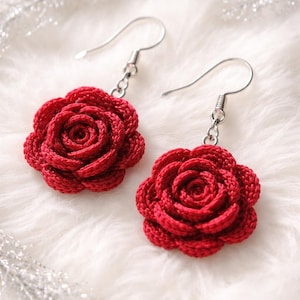 Puede incluir: Un par de pendientes de rosas rojas de ganchillo. Los pendientes presentan un diseño floral detallado con un centro en espiral y pétalos en capas. Cuelgan de ganchos plateados. El fondo es de un material suave, blanco y esponjoso.