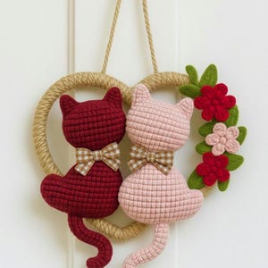 Op de afbeelding: Handgemaakte gehaakte kattenwanddecoratie. De katten zijn bordeauxrood en roze, elk met een geruite strik. Ze zijn geplaatst op een hartvormige krans van touw en versierd met bloemen.