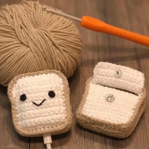 May include: Crocheted toast-shaped phone charger cases with smiling faces. The cases are white with tan edges, and one has a flap with a decorative button. A ball of tan yarn and an orange crochet hook are in the background.