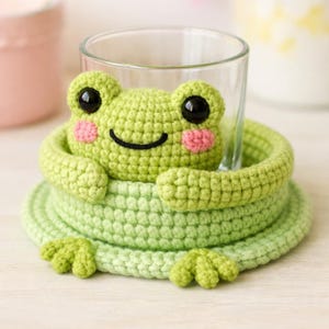 May include: A handmade, light green crochet frog coaster with a smiling face, pink cheeks, and black eyes. The coaster is designed to hold a glass, with the frog's head and arms encircling the glass. The coaster has a round base and frog feet.