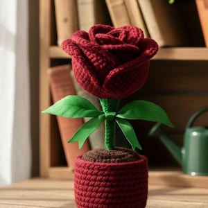 Puede incluir: Una rosa de ganchillo hecha a mano en una maceta roja. La rosa es de color burdeos intenso, con hojas verdes y un tallo verde. La maceta también es roja y la tierra es marrón. La rosa es un artículo decorativo.