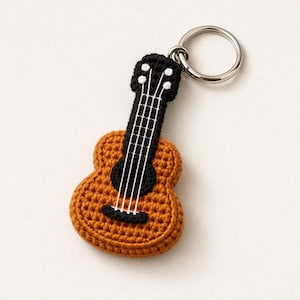 Könnte beinhalten: Ein gehäkelter Gitarren-Schlüsselanhänger in Braun-, Schwarz- und Weißtönen. Das Miniaturinstrument hat ein detailliertes Design mit Saiten und einem silbernen Schlüsselring. Das handgefertigte Accessoire ist ein charmantes Geschenk.
