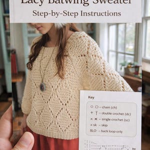 Puede incluir: Un libro de patrones de ganchillo titulado "Lacy Batwing Sweater" con instrucciones paso a paso. La portada presenta un suéter color crema con un patrón de diamantes. El libro incluye una clave de símbolos de ganchillo y un gráfico de símbolos Grechet.